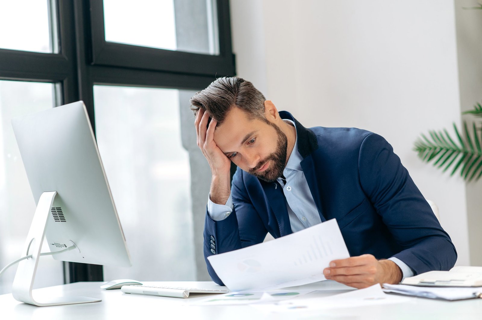 Ein Mann im Business-Outfit sitzt nachdenklich am Schreibtisch und betrachtet Unterlagen – symbolisch für wirtschaftliche Belastungen und steigende Unternehmensausgaben.